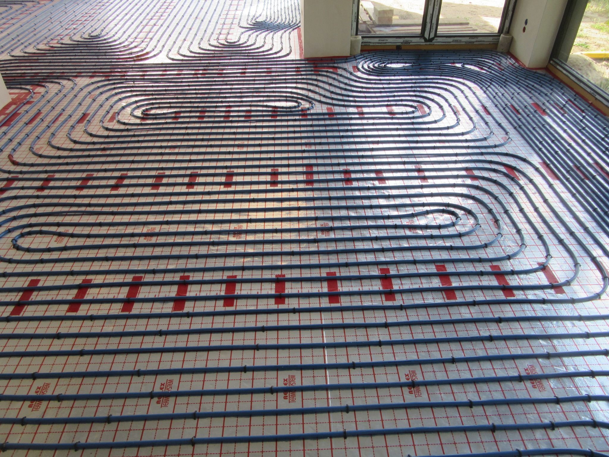 Ułożone, niebieskie rury ogrzewania podłogowego na folii izolacyjnej z czerwonym nadrukiem kratki, widoczne okno i fragment ściany.