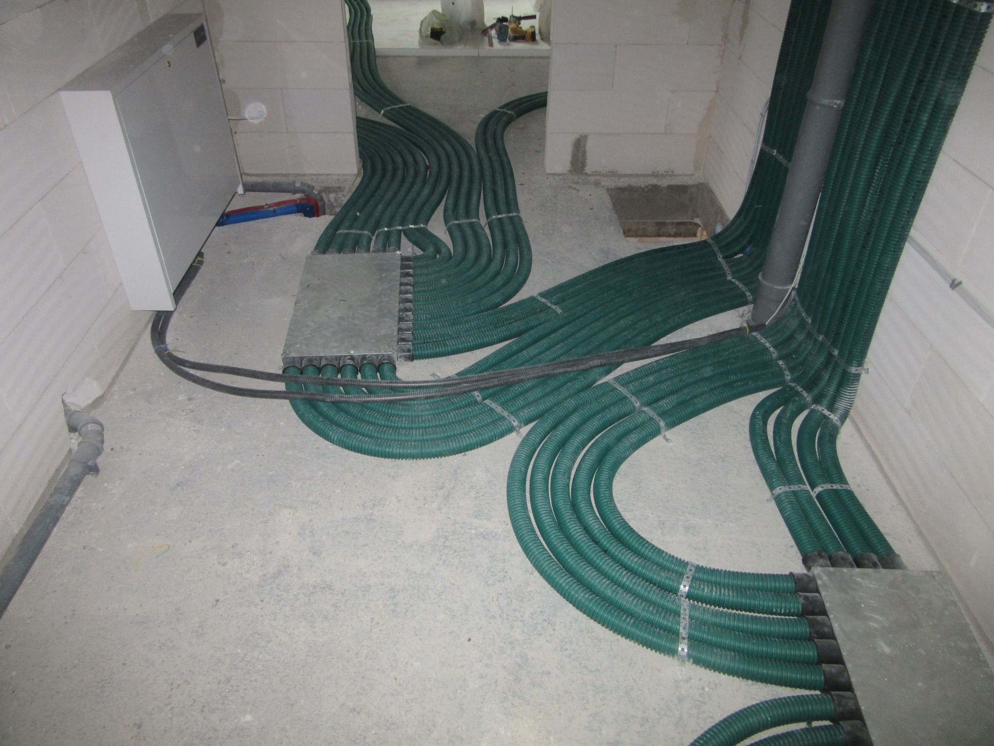 Zielone, karbowane rury instalacji w podłodze, prowadzone w peszlach, z metalowymi puszkami rozdzielczymi, w surowym stanie wykończenia wnętrza.