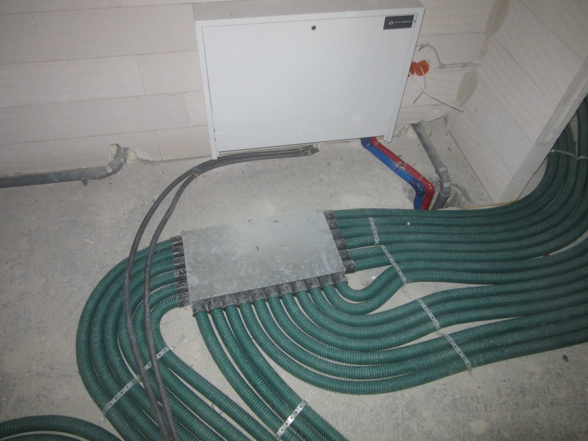 Rozdzielacz ogrzewania podłogowego z zielonymi rurami w surowym wnętrzu. Widoczna skrzynka rozdzielcza na ścianie i fragment instalacji wodnej z rurami czerwono-niebieskimi.