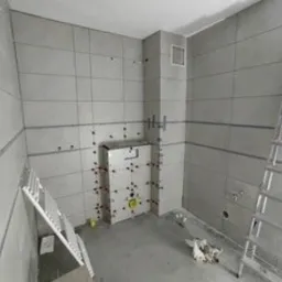 Wnętrze łazienki w trakcie wykańczania, z szarymi, prostokątnymi płytkami na ścianach i częściowo obudowaną toaletą podtynkową. Widoczne drabiny i narzędzia glazurnicze.