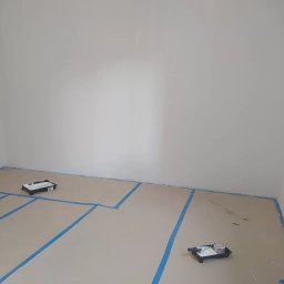 NaPoziomie - Pokój przygotowany do malowania: zabezpieczona podłoga kartonem i taśmą, widoczne kuwety malarskie. Świeżo pomalowana ściana w jasnym odcieniu.