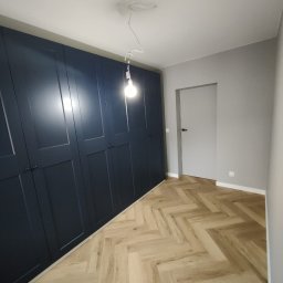 NaPoziomie - Granatowa szafa wnękowa z ciemnymi uchwytami, parkiet w jodełkę i szara ściana z białymi listwami przypodłogowymi. Widoczna żarówka na kablu.