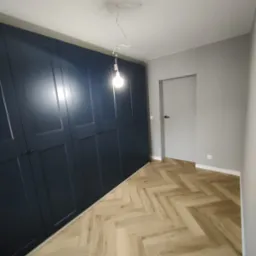 Granatowa szafa wnękowa z ciemnymi uchwytami, parkiet w jodełkę i szara ściana z białymi listwami przypodłogowymi. Widoczna żarówka na kablu.