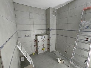 Wnętrze łazienki w trakcie wykańczania, z szarymi, prostokątnymi płytkami na ścianach i częściowo obudowaną toaletą podtynkową. Widoczne drabiny i narzędzia glazurnicze.