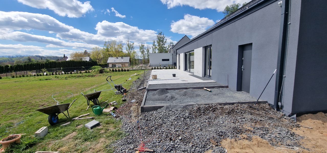 Układanie kostki brukowej na tarasie domu jednorodzinnego z szarą elewacją. Widoczne narzędzia, piasek i częściowo ułożona kostka. W tle trawnik i zabudowa.