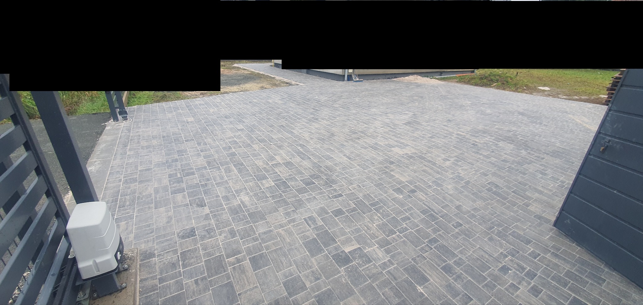 Ułożona szara kostka brukowa na podjeździe, widok z góry, z fragmentem bramy w tle. Automatyka do bramy widoczna po lewej stronie.