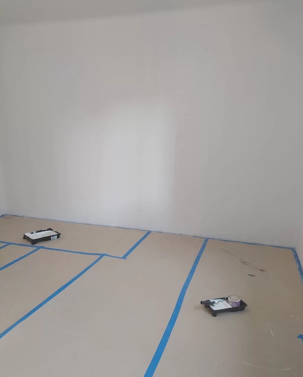 Pokój przygotowany do malowania: zabezpieczona podłoga kartonem i taśmą, widoczne kuwety malarskie. Świeżo pomalowana ściana w jasnym odcieniu.