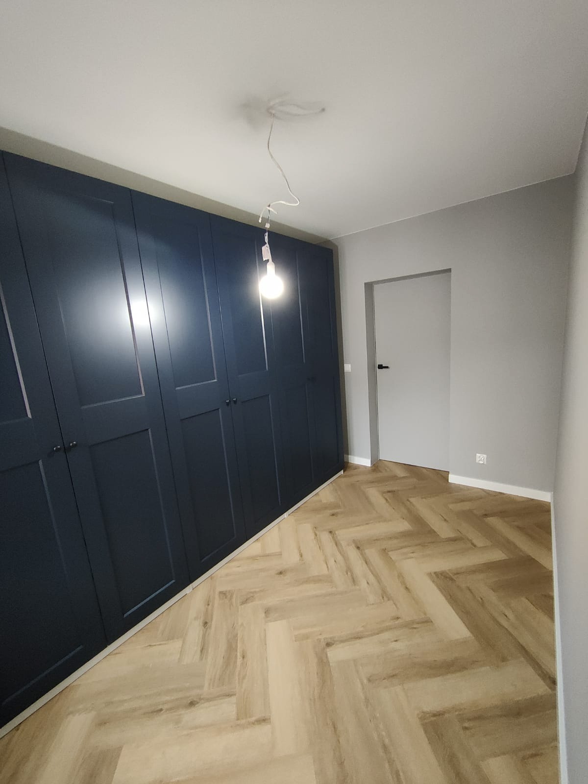 Granatowa szafa wnękowa z ciemnymi uchwytami, parkiet w jodełkę i szara ściana z białymi listwami przypodłogowymi. Widoczna żarówka na kablu.