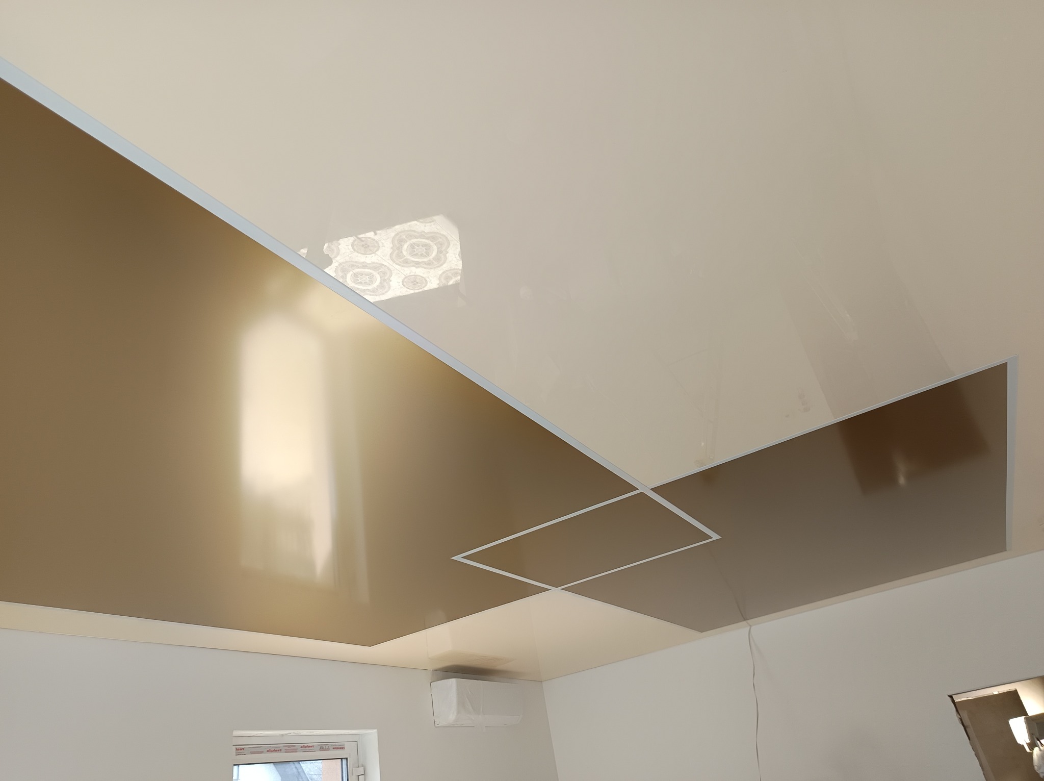 Beżowo-złoty sufit napinany z geometrycznym wzorem, widoczny z dołu. Fragment okna i klimatyzacji w tle. Połyskująca powierzchnia odbija światło.