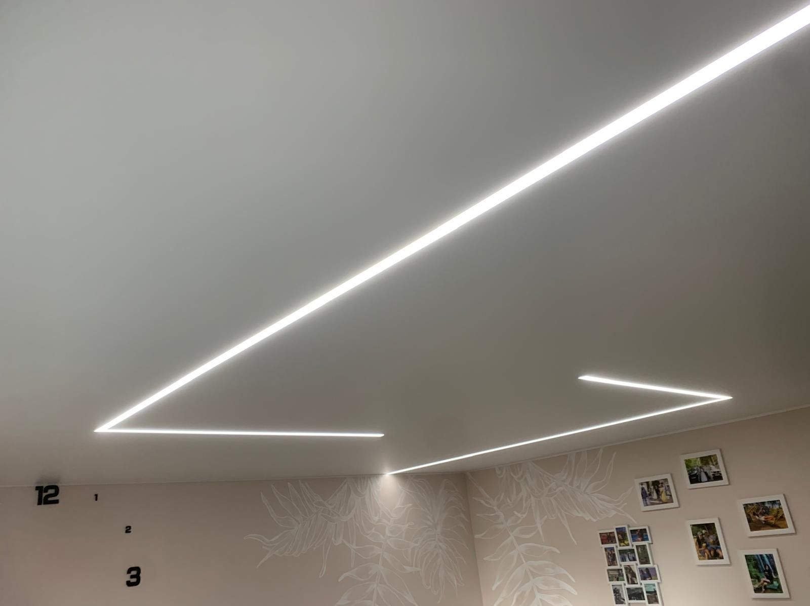 Jasny, biały sufit napinany z geometrycznym wzorem z linii LED. Ściany z motywem roślinnym i ramkami zdjęć. Nowoczesne oświetlenie wnętrza.