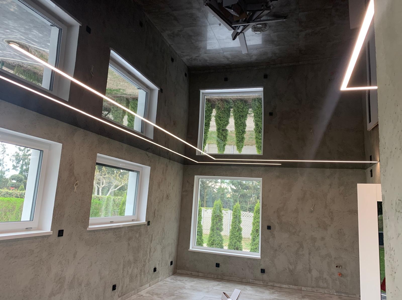 Nowoczesne wnętrze z szarymi ścianami o fakturze, białymi oknami i liniowym oświetleniem LED. Widok na zieleń za oknami, sufit wykończony płytkami w odcieniach szarości.