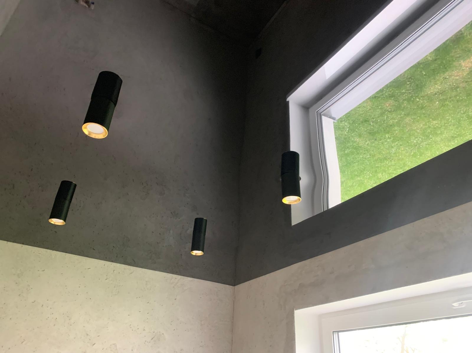 Nowoczesne oświetlenie sufitowe z trzema czarnymi lampami tubowymi, szary sufit z efektem betonu, okno z widokiem na zieleń. Minimalistyczne wykończenie wnętrza.