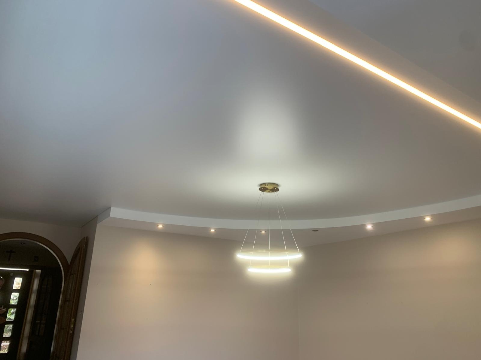 Biały sufit napinany z oświetleniem LED liniowym i okrągłą lampą wiszącą. Widoczne punkty świetlne wzdłuż zakrzywionej krawędzi. Minimalistyczny design.