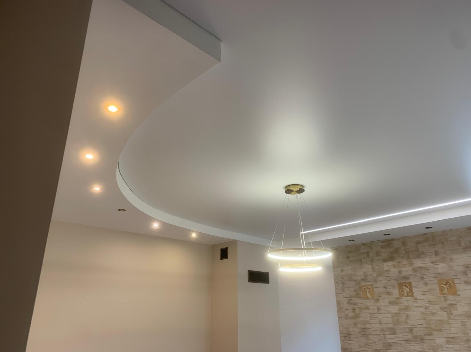 Biały, podwieszany sufit z zaokrąglonymi krawędziami, oświetlony punktowo i lampą wiszącą. Ściana z dekoracyjną okładziną kamienną.