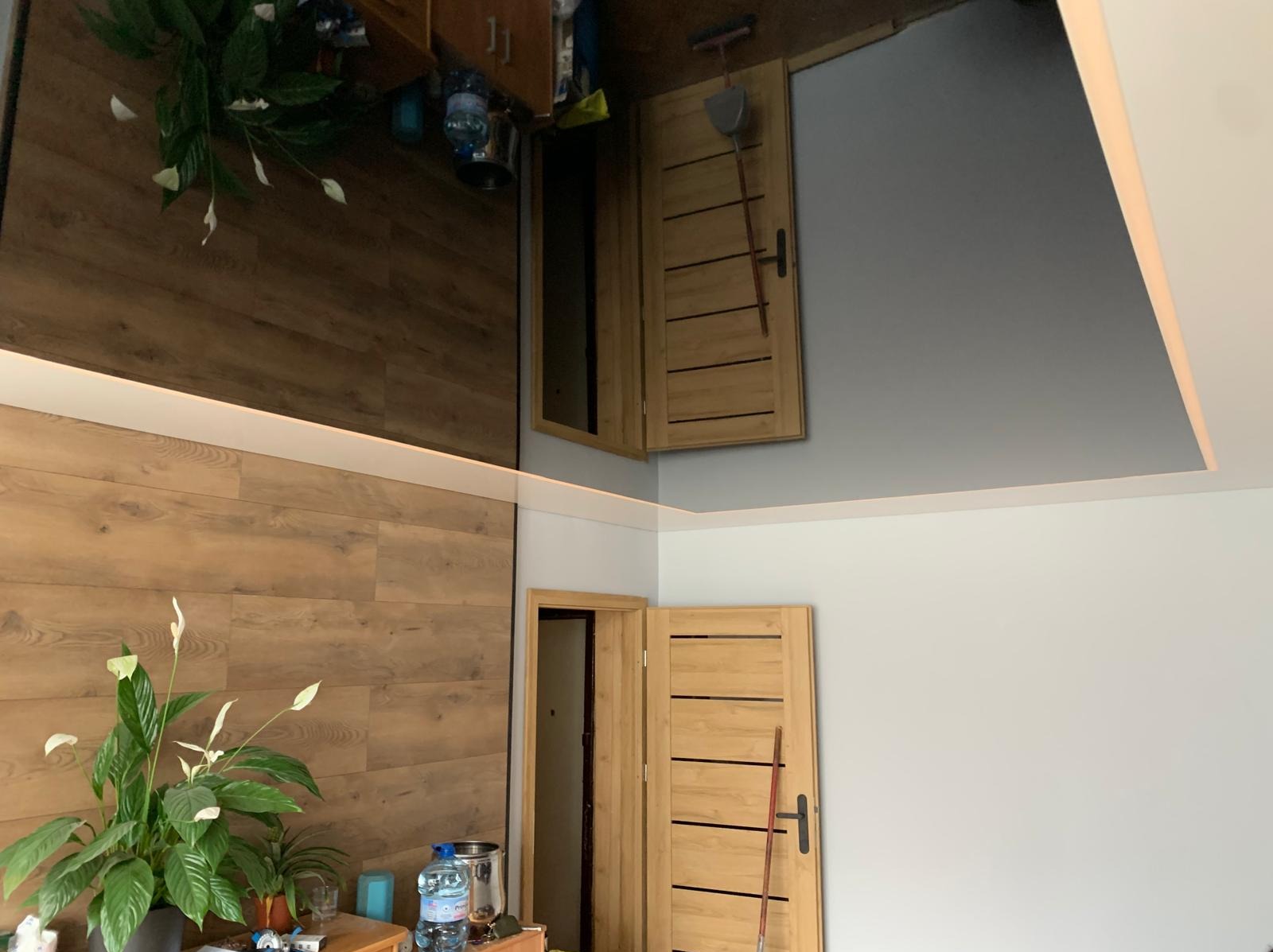 Nowoczesne wnętrze z drewnianą ścianą i lustrzanym sufitem, odbijającym fragment pomieszczenia. Widoczna drewniana stolarka drzwiowa i oświetlenie LED.