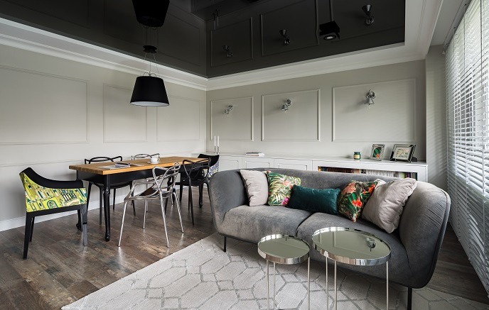 Eleganckie wnętrze: szary narożnik z poduszkami, stoliki kawowe, stół z krzesłami i błyszczący, ciemny sufit. Całość w stonowanej kolorystyce.