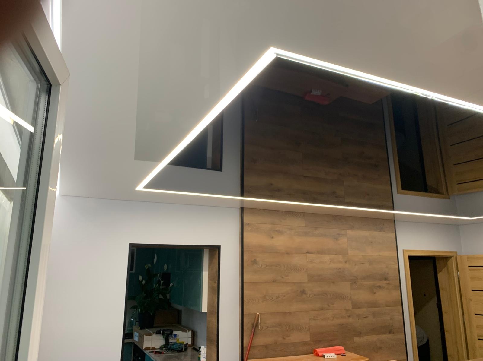 Biały, błyszczący sufit napinany z oświetleniem LED w kształcie litery L, odbijający fragment drewnianej ściany i okna, widok z dołu.