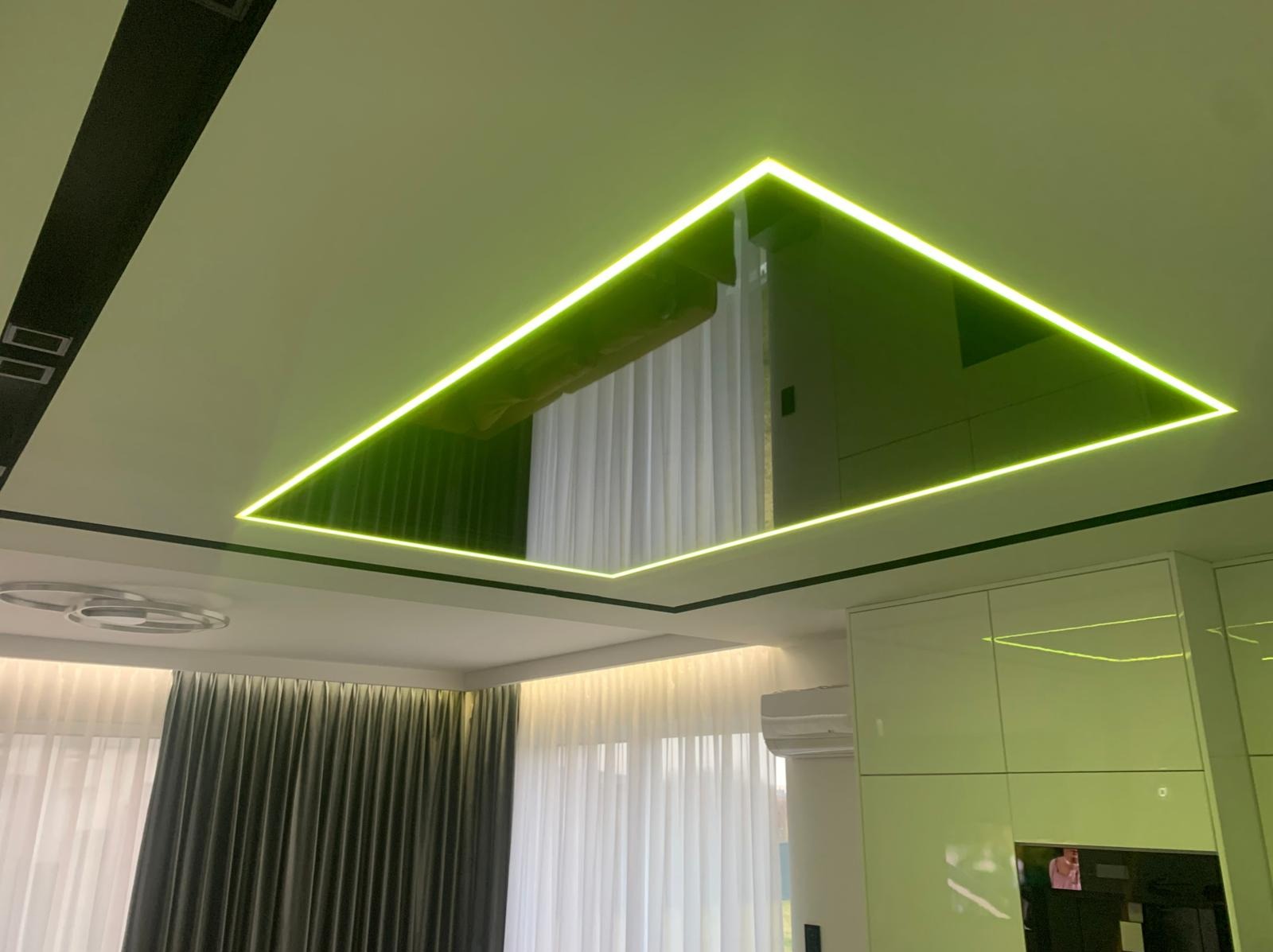 Nowoczesny sufit napinany z podświetleniem LED w kształcie trapezu, odbijający wnętrze pokoju. Widoczne zasłony i fragmenty mebli w odbiciu.