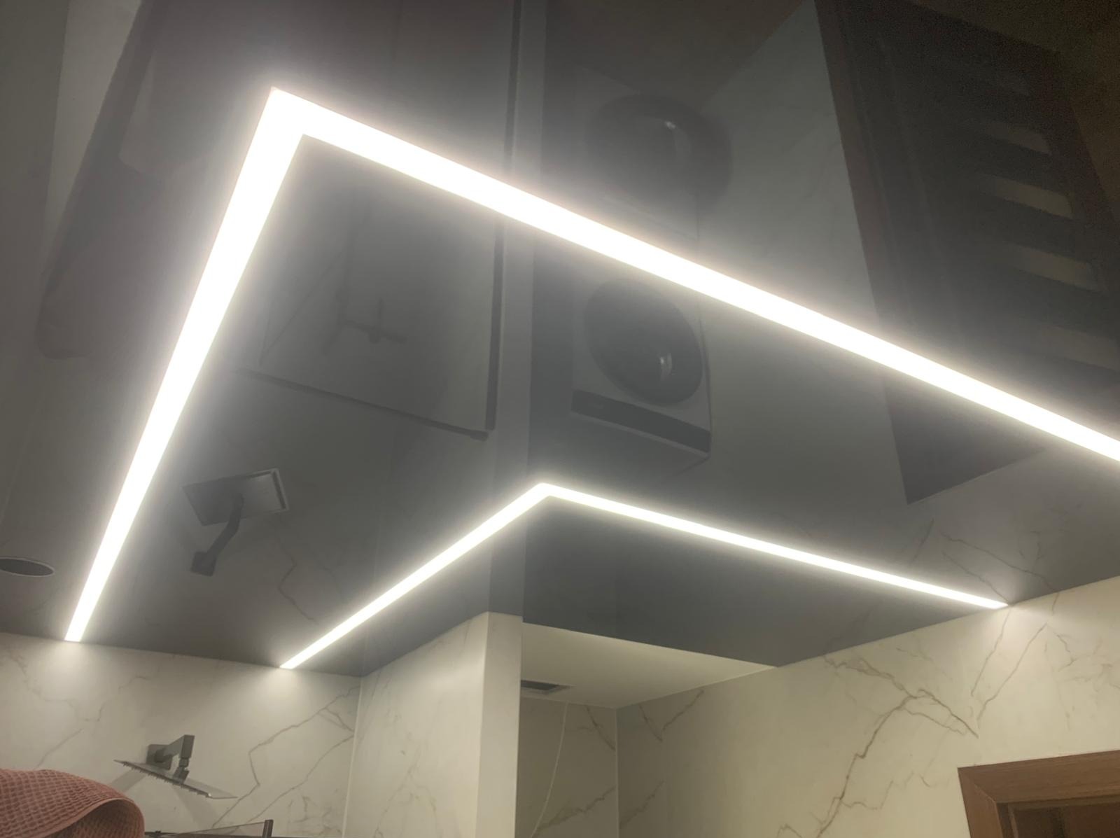 Błyszczący, grafitowy sufit z liniowym oświetleniem LED w kształcie litery L, odbijający elementy łazienki, w tym armaturę i marmurowe płytki.