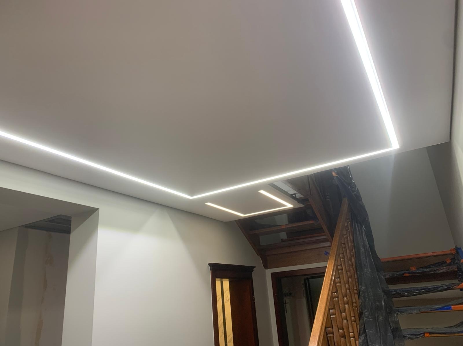 Nowoczesny sufit z liniowym oświetleniem LED w kształcie kwadratów, drewniane schody w tle. Wykończenie wnętrza w odcieniach bieli i drewna.