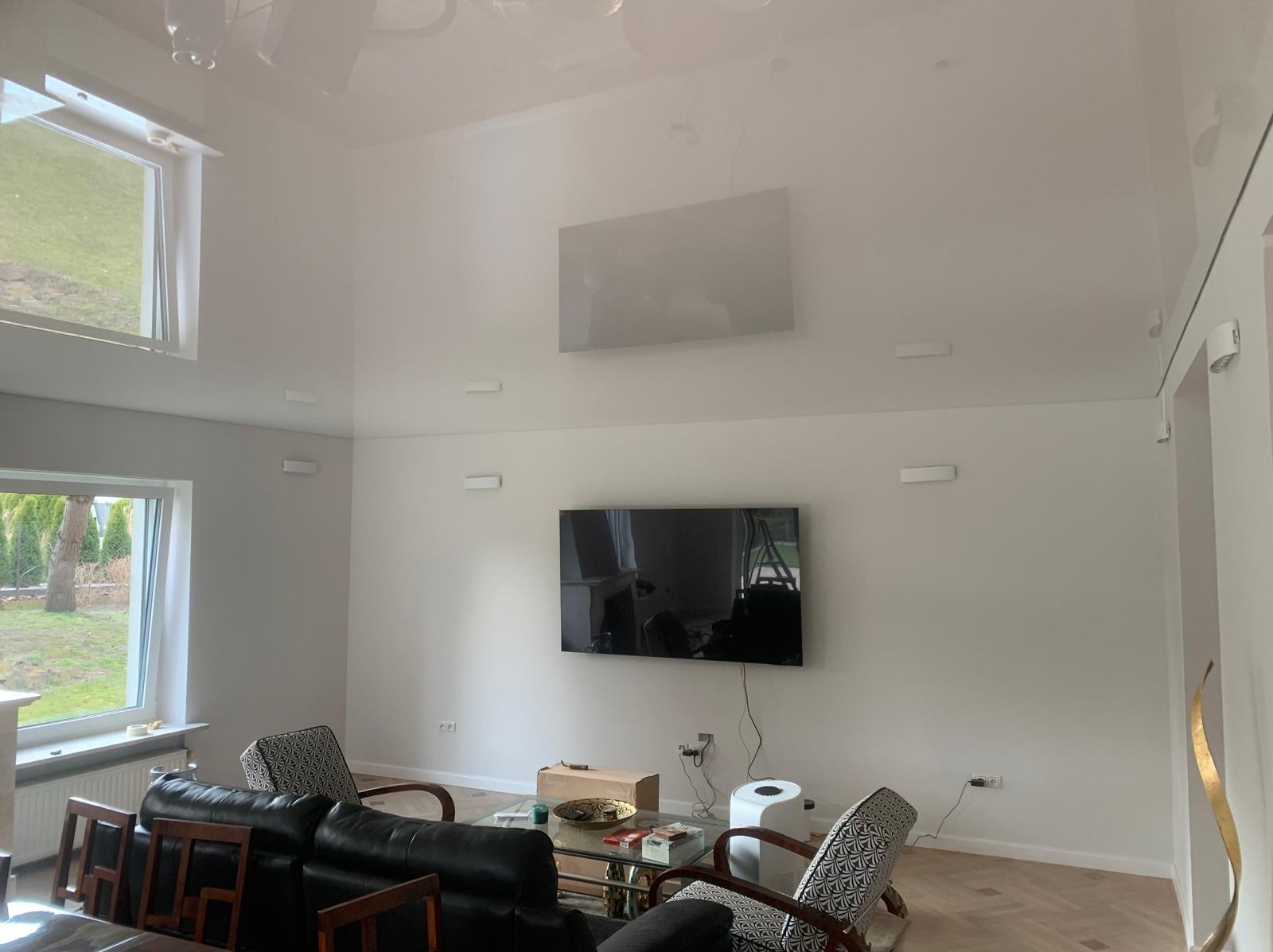 Biały salon z połyskującym sufitem napinanym, oświetleniem punktowym i telewizorem na ścianie. Widoczne meble i okna z widokiem na zieleń.