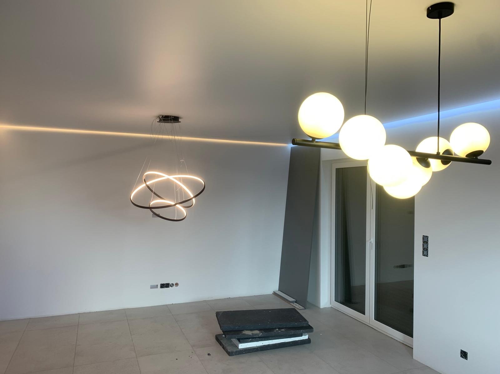 Nowoczesne oświetlenie w salonie: lampa pierścieniowa i listwa LED na suficie napinanym, kuliste lampy wiszące, jasne ściany i płytki podłogowe, minimalistyczny design.