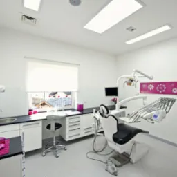 Jasne wnętrze gabinetu lekarskiego z nowoczesnym fotelem dentystycznym, umywalką i oknem z widokiem na domy. Całość utrzymana w bieli z akcentami różu.