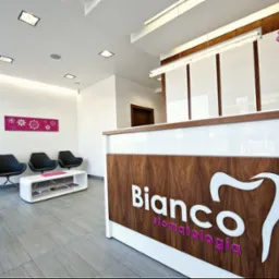 Nowoczesna recepcja kliniki stomatologicznej z logo 'Bianco stomatologia', kwiatami i designerskimi krzesłami w poczekalni.