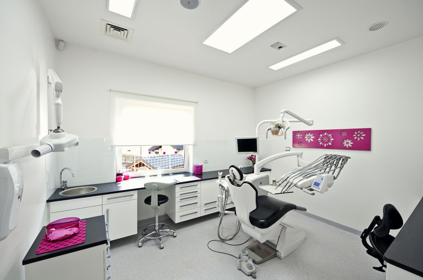 Jasne wnętrze gabinetu lekarskiego z nowoczesnym fotelem dentystycznym, umywalką i oknem z widokiem na domy. Całość utrzymana w bieli z akcentami różu.