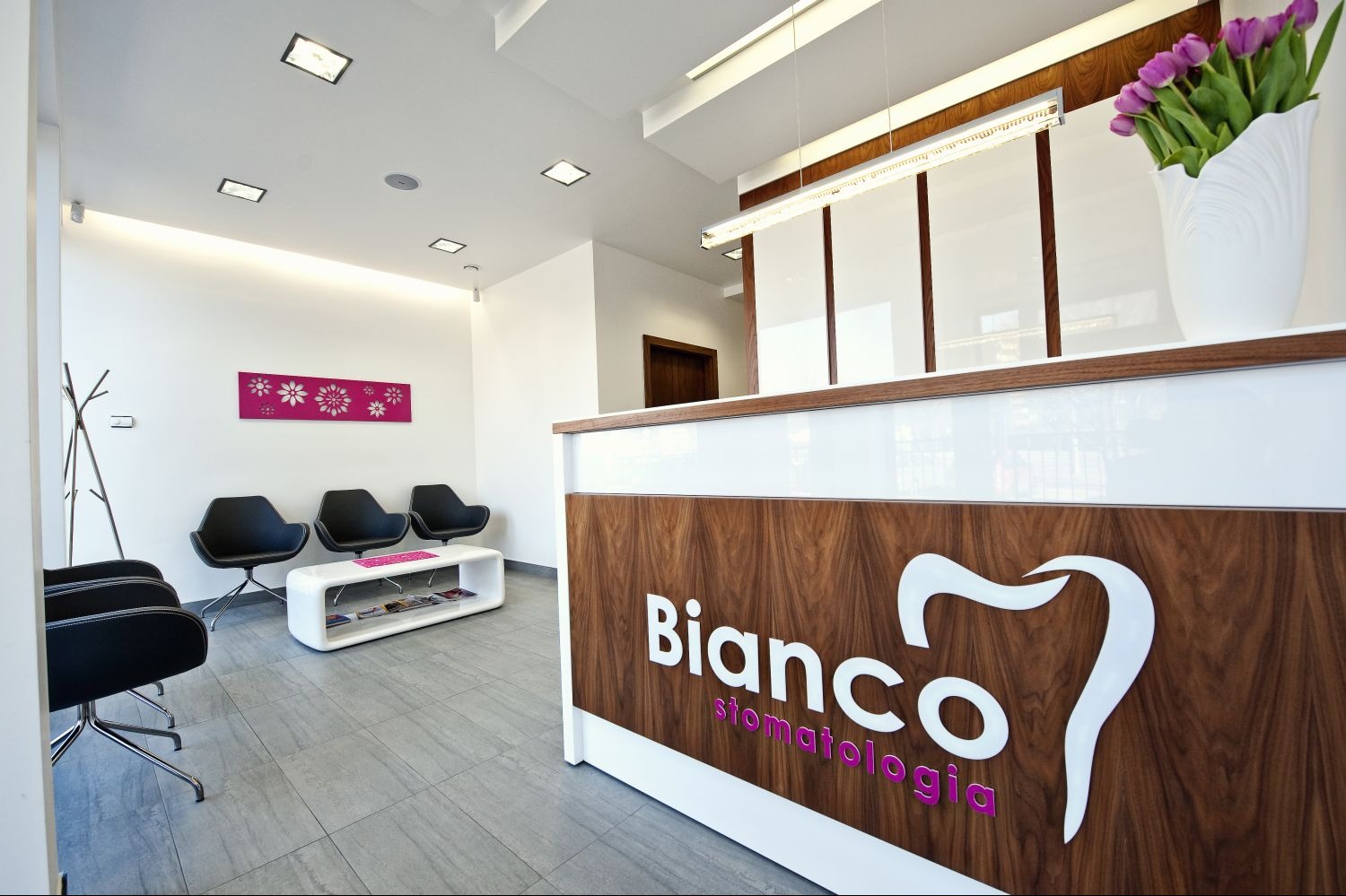 Nowoczesna recepcja kliniki stomatologicznej z logo 'Bianco stomatologia', kwiatami i designerskimi krzesłami w poczekalni.