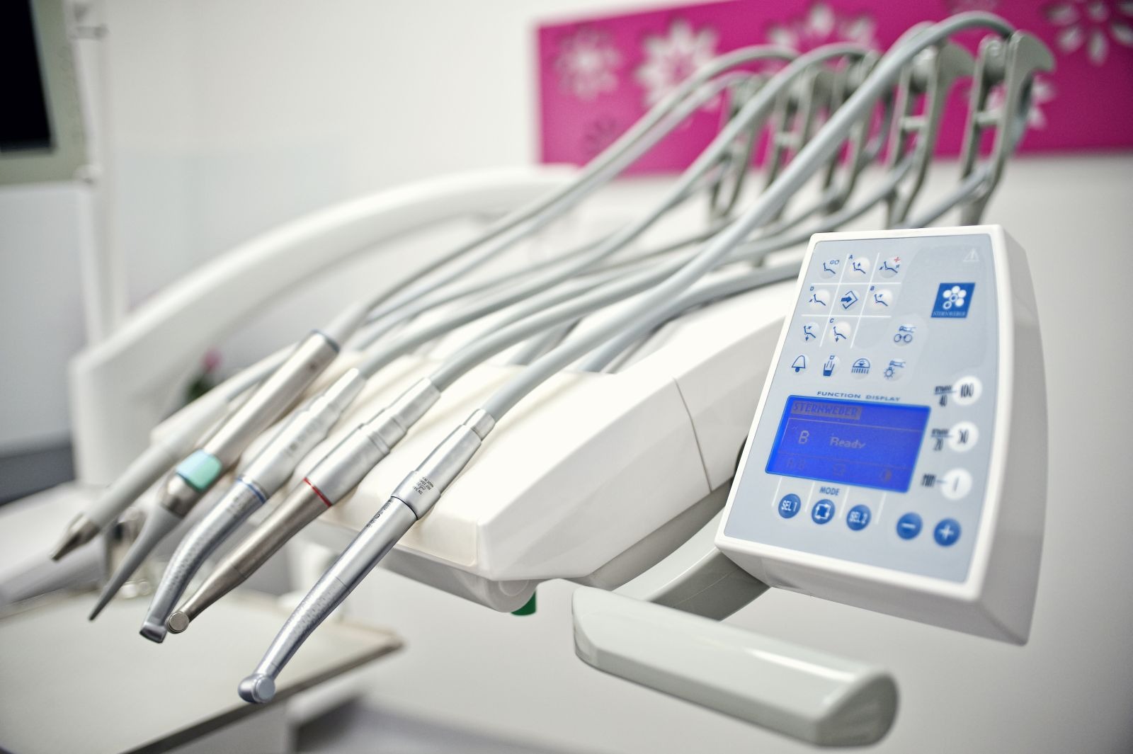 Nowoczesny sprzęt dentystyczny z panelem sterowania i końcówkami w zbliżeniu, na tle ściany w gabinecie. Widoczne detale i ergonomiczny design.