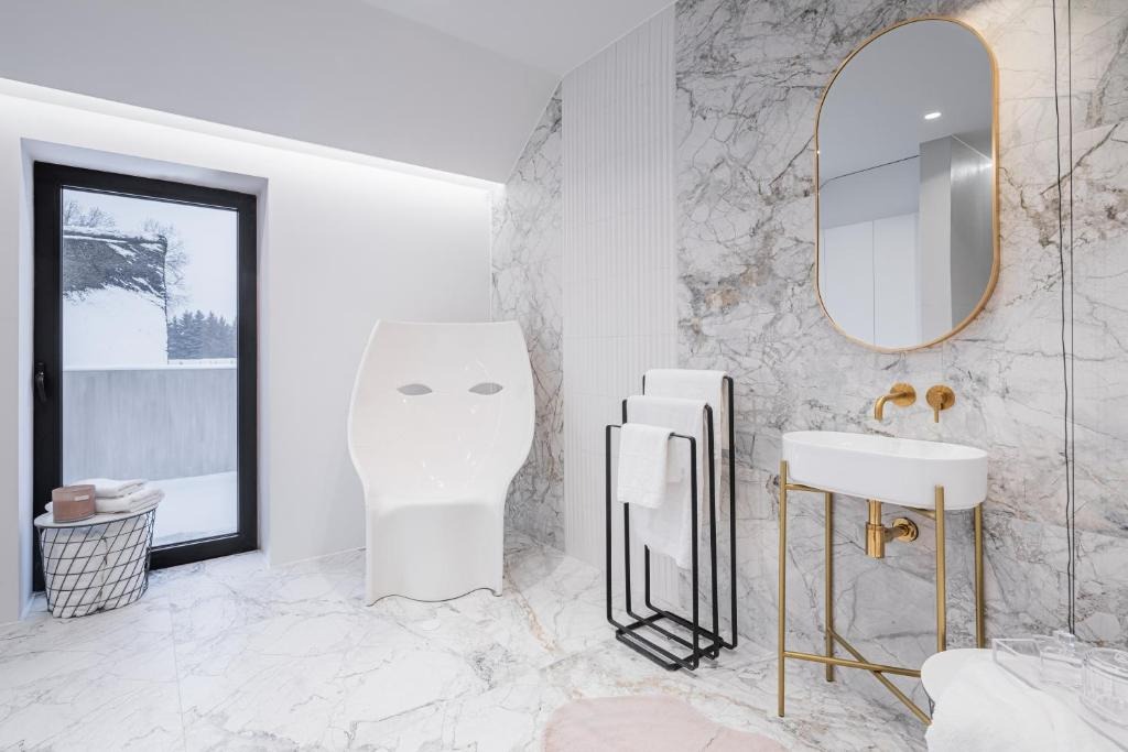 Stylowa łazienka z marmurowymi płytkami, owalnym lustrem w złotej ramie, białą umywalką na złotym stelażu i nowoczesnym krzesłem. Za oknem zimowy krajobraz.