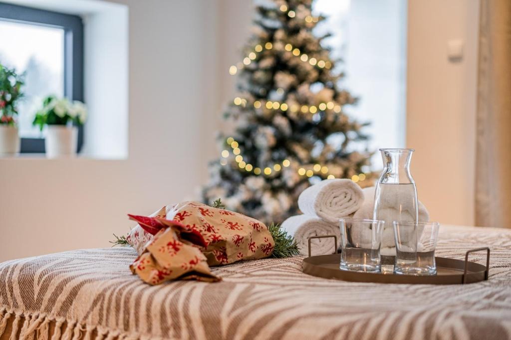 Świąteczny wystrój pokoju na wynajem: prezenty, ręczniki, woda i choinka z lampkami tworzą przytulną atmosferę. Okno z kwiatami w tle.