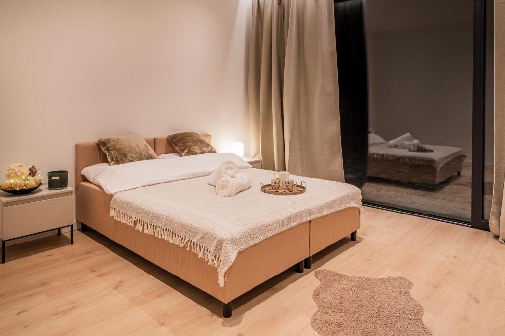 Przytulna sypialnia z beżowym łóżkiem, miękkimi poduszkami i dekoracyjnym pledem. Na łóżku ręczniki i taca z napojami. Widok na taras przez duże okno.