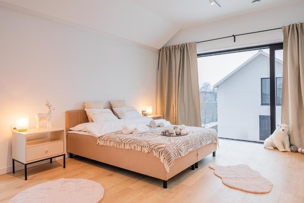 Jasna sypialnia z beżowym łóżkiem, dekoracjami i dużym oknem z widokiem na ośnieżony krajobraz. Na podłodze drewniany parkiet i okrągły dywanik.