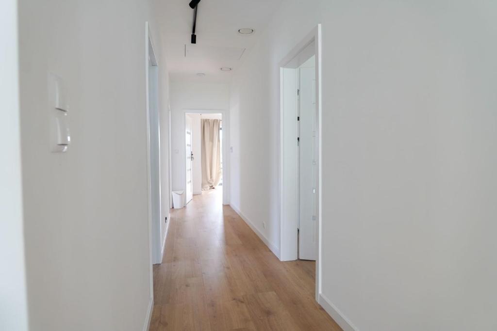 Jasny korytarz z drewnianą podłogą i białymi ścianami. Widoczne drzwi i oświetlenie sufitowe. Minimalistyczny design wnętrza, perspektywa w głąb mieszkania.