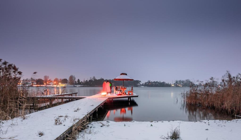 Zimowy krajobraz: ośnieżony pomost nad jeziorem z miejscem do siedzenia pod parasolem i ogniskiem. Romantyczna sceneria o zmierzchu.