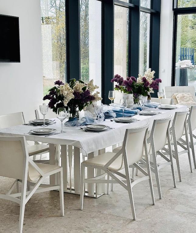 Elegancko nakryty stół z białym obrusem, zastawą i kwiatami w wazonach, ustawiony w jasnym pomieszczeniu z dużymi oknami, sugerujący przygotowanie do uroczystości.