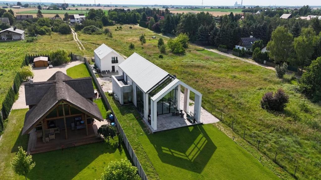Nowoczesny dom z dużymi przeszkleniami i białym dachem sąsiaduje z tradycyjnym domem krytym strzechą. Widok z lotu ptaka na zadbany teren.