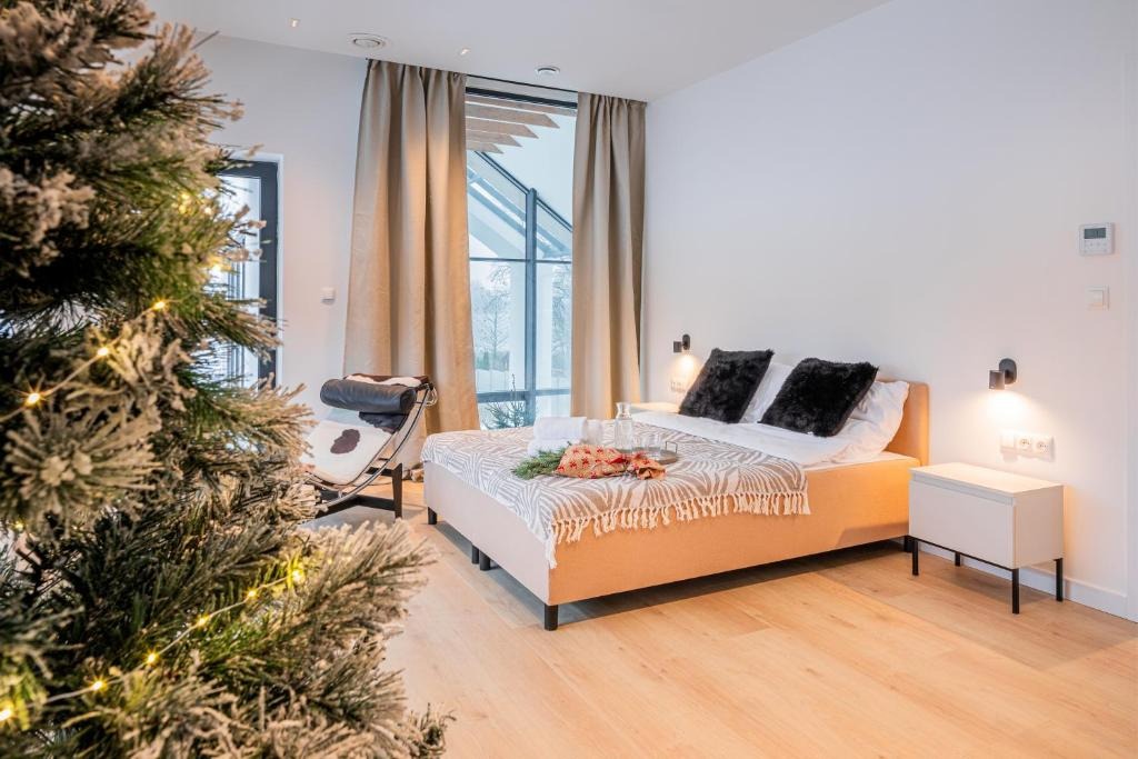 Nowoczesna sypialnia z łóżkiem, stolikiem nocnym i fotelem Barcelona. Świąteczny akcent: choinka z lampkami. Duże okno z widokiem na zewnątrz. Jasne drewniane podłogi.