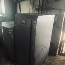 Kocioł na pellet Kolton Bio Pellmax w kotłowni. Widoczne rury instalacji grzewczej i ściany z surowego betonu. Na kotle wyświetlacz i oznaczenia modelu.