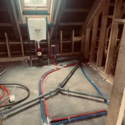 Instalacja wodna w poddaszu z oknem dachowym: rury w izolacji czerwonej i niebieskiej, szara kanalizacja, stelaż podtynkowy Grohe. Widoczne elementy konstrukcji drewnianej.