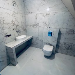 Hydro-klim - Wykończona łazienka w bieli i marmurze: umywalka nablatowa, toaleta podwieszana z niebieskim przyciskiem, płytki imitujące marmur na ścianach i podłodze.