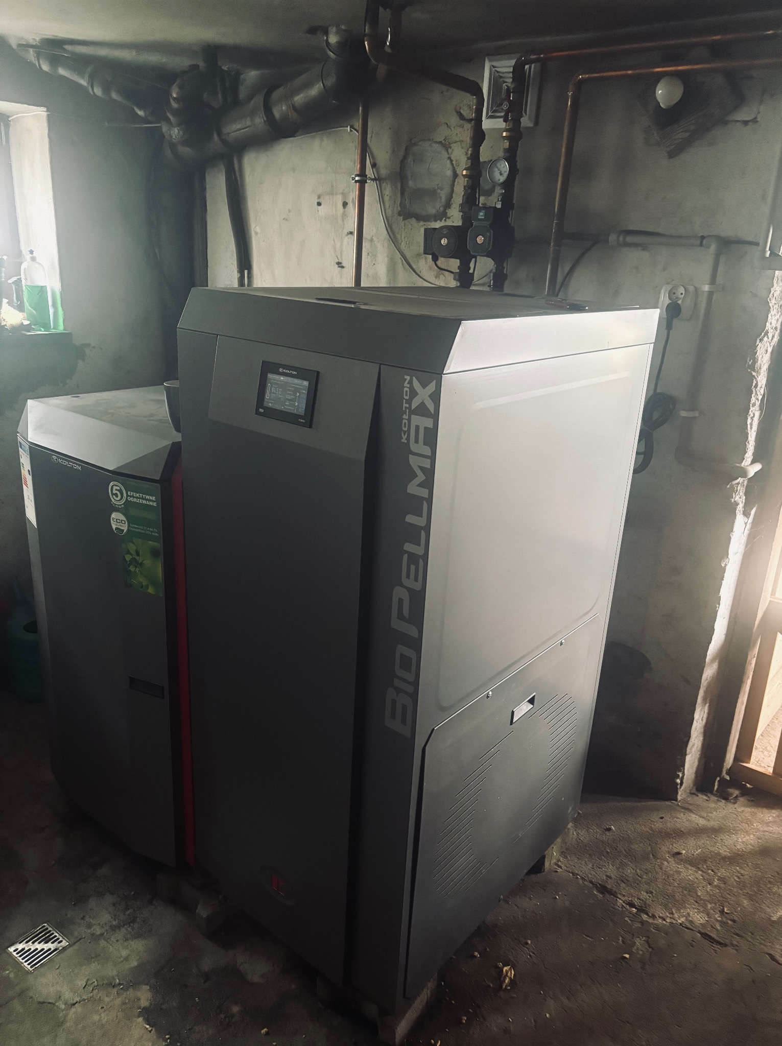Kocioł na pellet Kolton Bio Pellmax w kotłowni. Widoczne rury instalacji grzewczej i ściany z surowego betonu. Na kotle wyświetlacz i oznaczenia modelu.