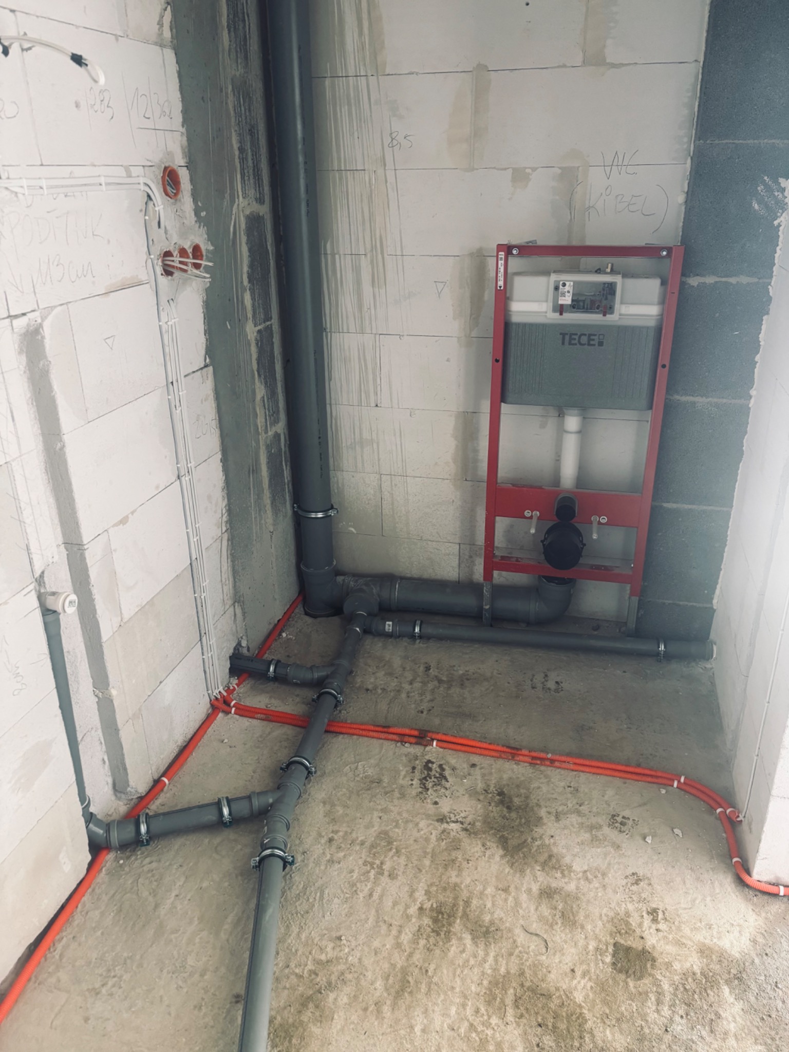 Surowa łazienka w trakcie instalacji: stelaż WC Tece, szare rury kanalizacyjne i pomarańczowe peszle instalacji wodnej, widoczne ślady po ołówku na ścianach.