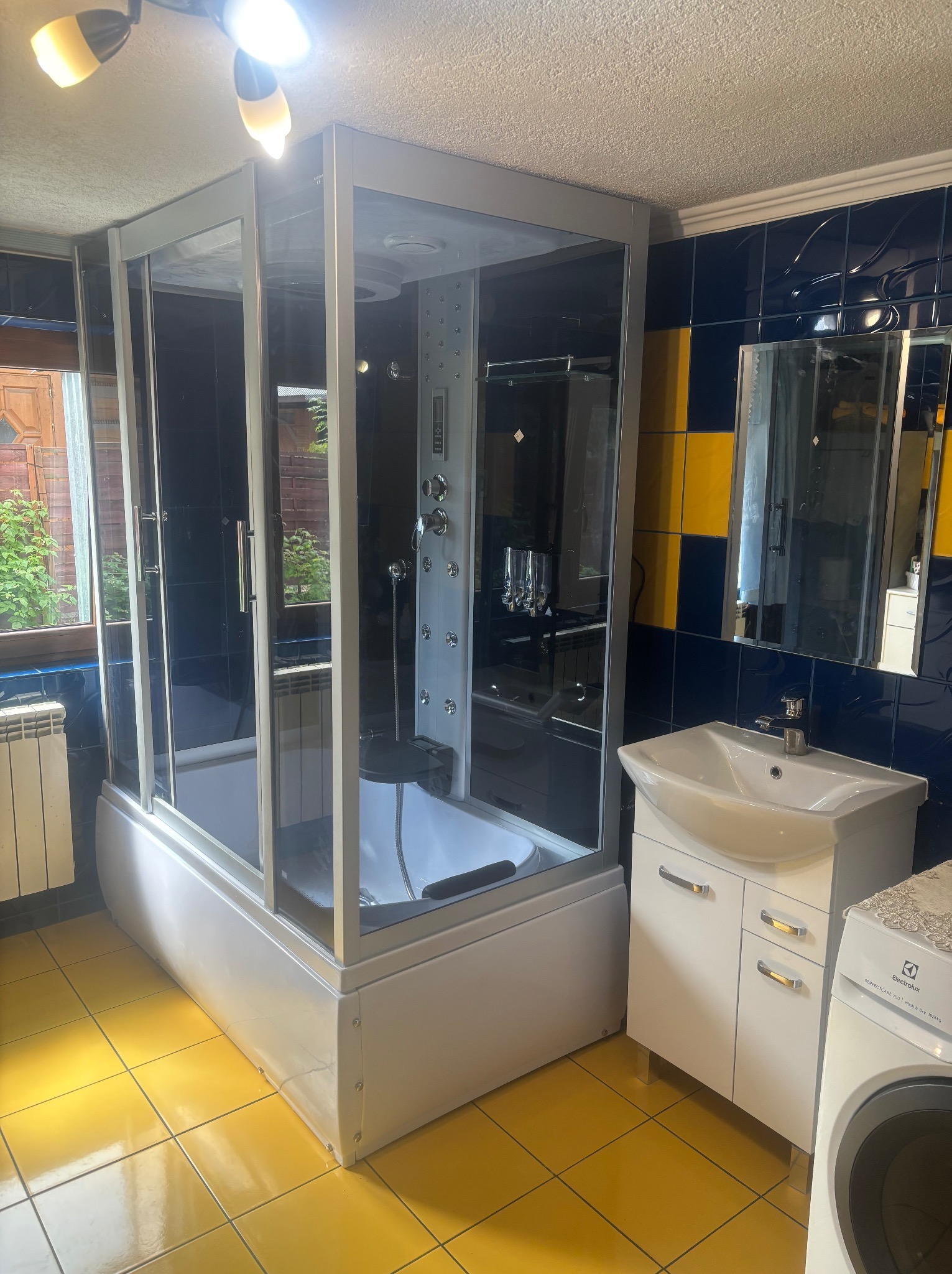 Kompletna kabina prysznicowa z hydromasażem w łazience z żółtą podłogą i ciemnymi płytkami. Widoczna umywalka z szafką i pralka. Okno z widokiem na zewnątrz.