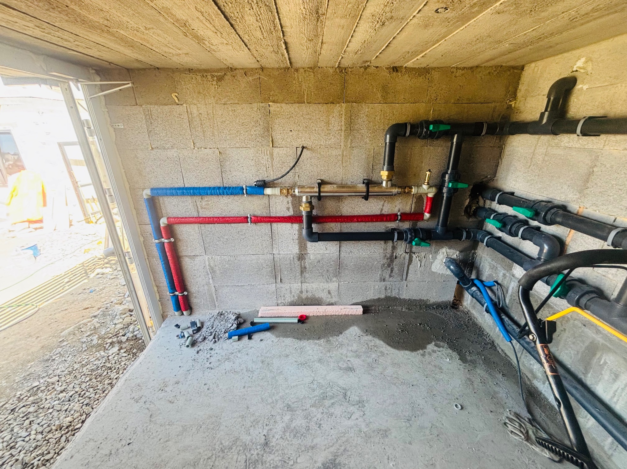 Węzeł instalacji wodnej z rurami w kolorach: niebieskim, czerwonym i czarnym,  z zielonymi zaworami, zamontowany w surowym pomieszczeniu z betonową podłogą.