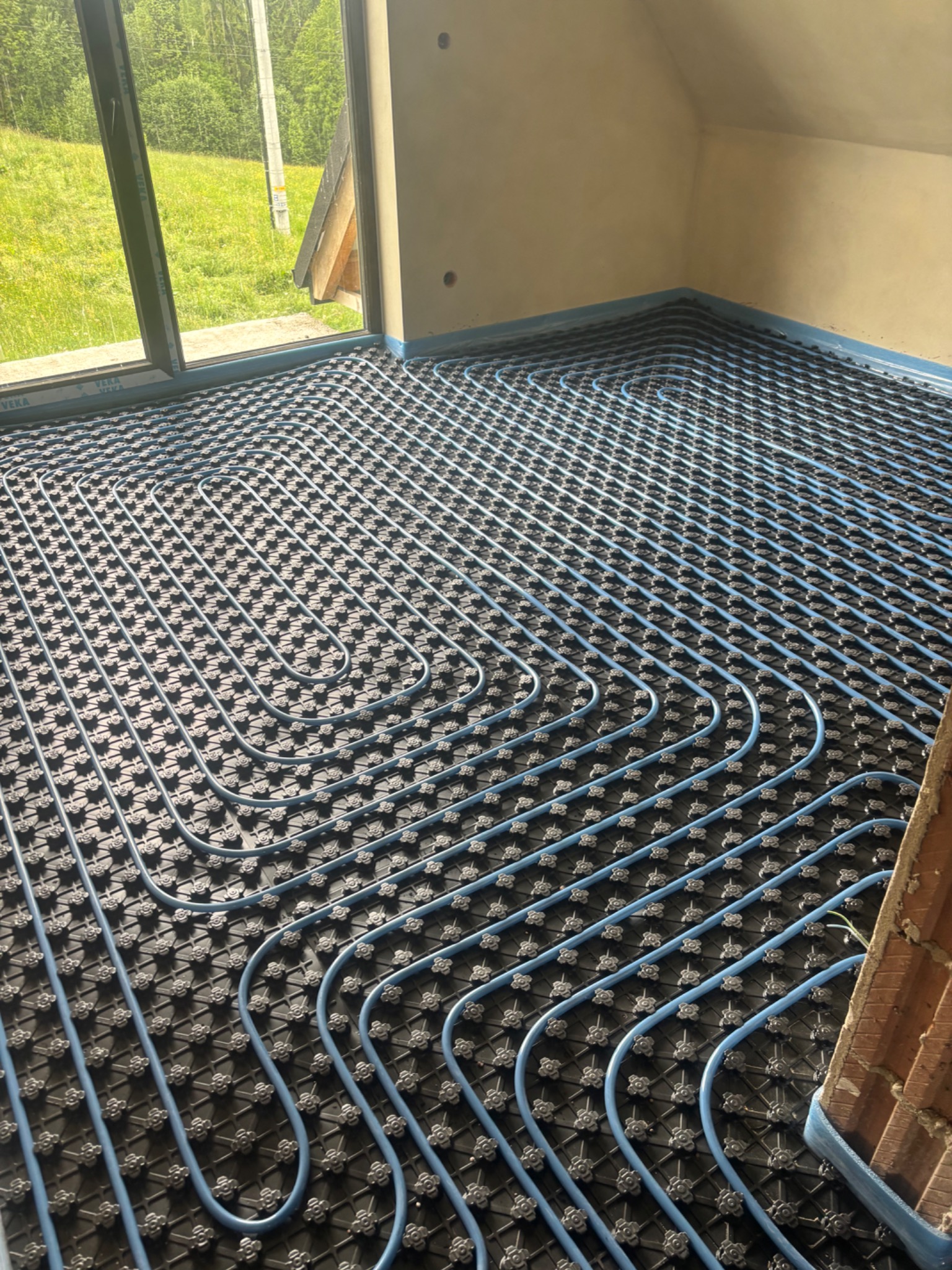 Niebieskie rury ogrzewania podłogowego ułożone na czarnej macie w pomieszczeniu z oknem i skośnym sufitem, widok z góry.