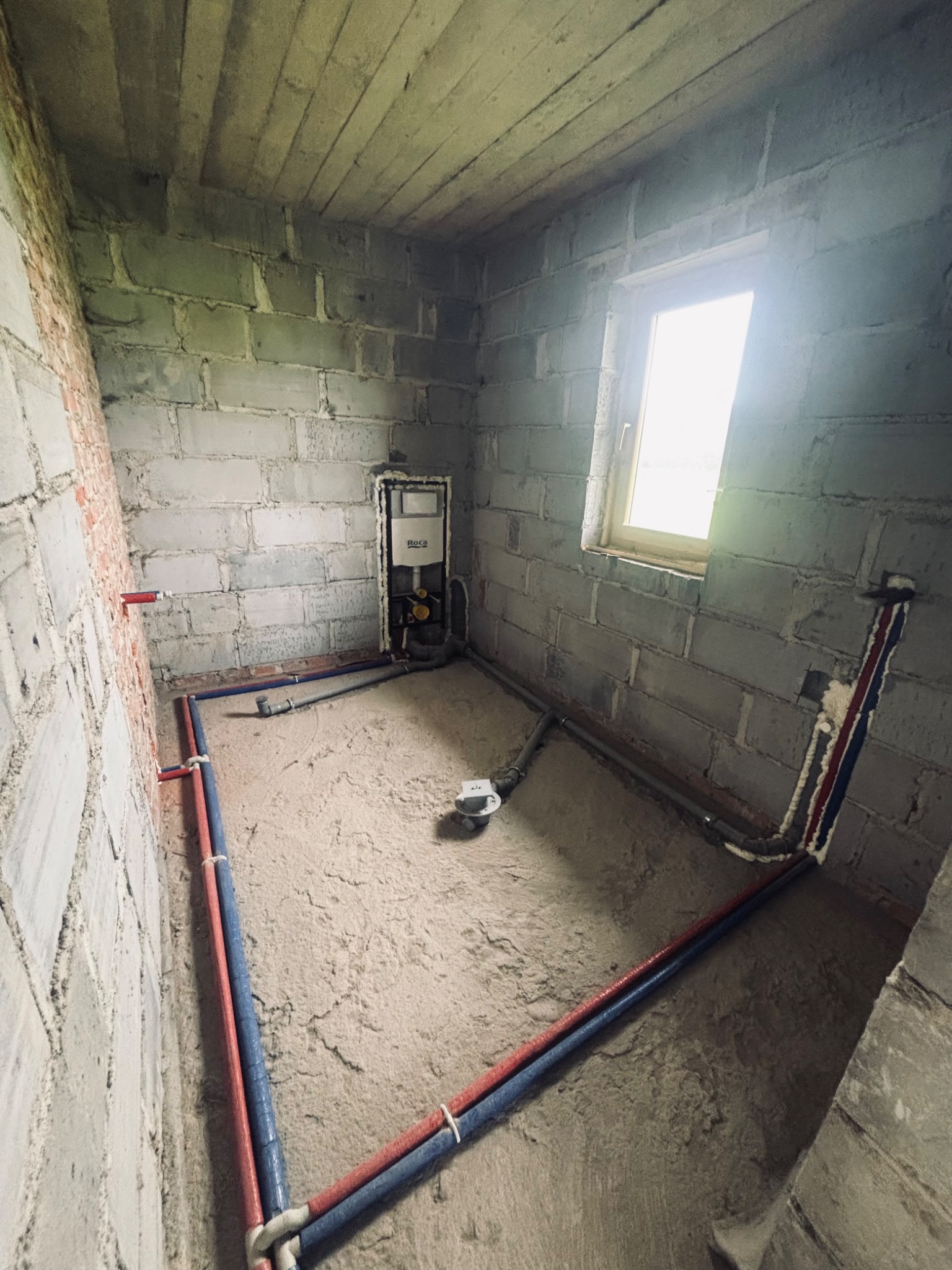 Surowy stan łazienki z częściowo rozprowadzoną instalacją wodno-kanalizacyjną z rur czerwonych i niebieskich. Widoczny stelaż podtynkowy WC i surowe ściany z betonu komórkowego.