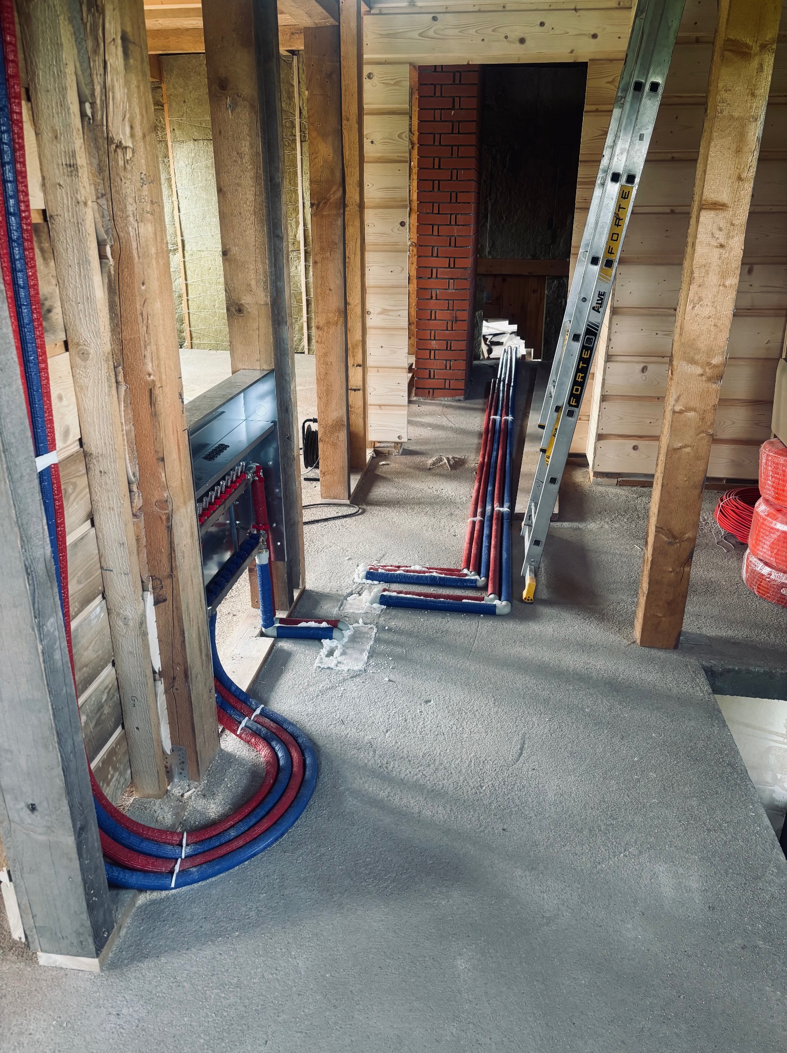 Instalacja ogrzewania podłogowego w surowym wnętrzu, rury w izolacji czerwono-niebieskiej, rozdzielacz w metalowej obudowie, drabina oparta o drewnianą konstrukcję.